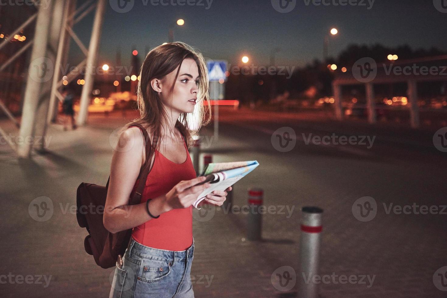 ung flicka kostar på natten nära terminalen på flygplatsen eller stationen och läsa stadskarta och letar efter hotell. söt turist med ryggsäckar bestämmer begreppet resa foto
