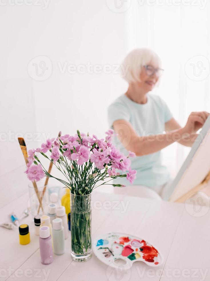 senior glad kvinna konstnär i glasögon med grått hår måla blommor i vas. kreativitet, konst, hobby, yrke koncept foto