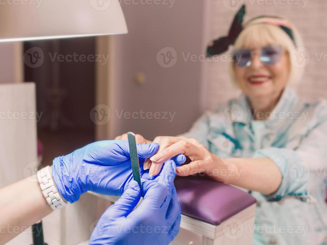 manikyrmästare i blåa handskar som krämer händerna på äldre snygg kvinna i blå solglasögon och jeansjacka som sitter på manikyrsalongen foto