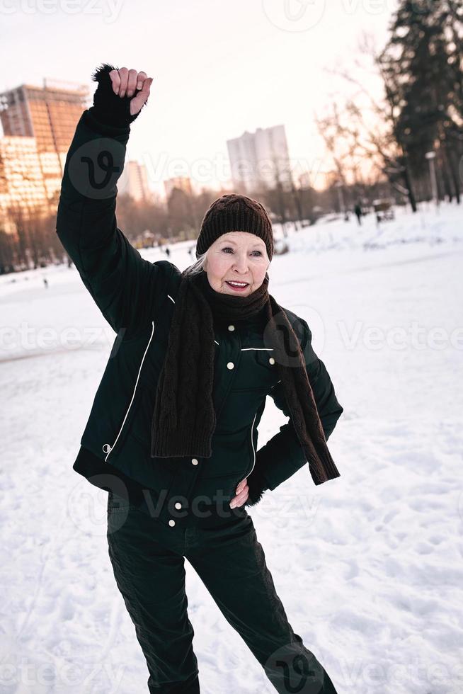 senior kvinna i hatt och sportig jacka gör sportövningar i snö vinterpark. vinter, ålder, sport, aktivitet, säsong koncept foto