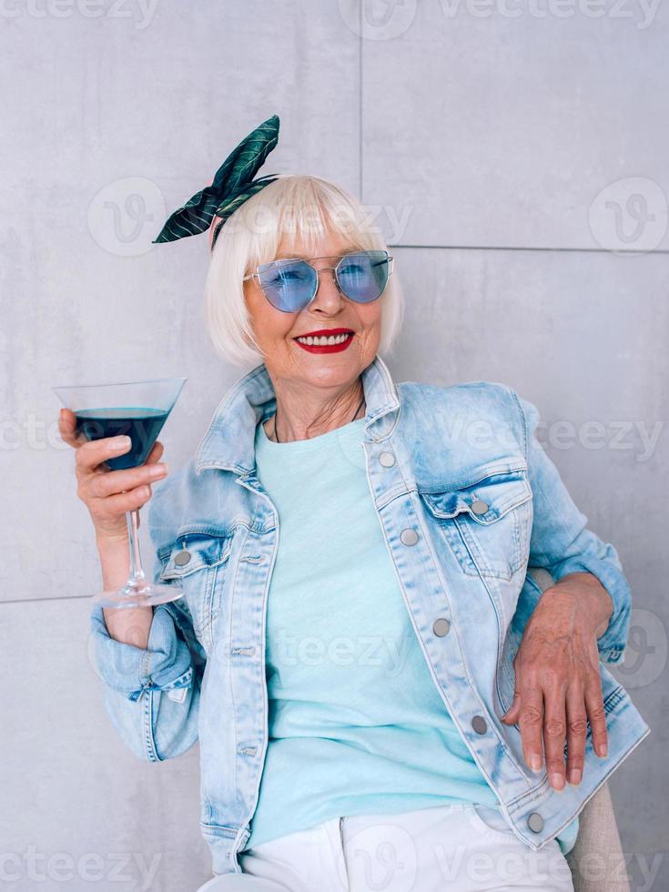 senior snygg kvinna med grått hår och i blå glasögon och jeansjacka håller glas med blå cocktail. alkohol, koppla av, semester, pension koncept foto