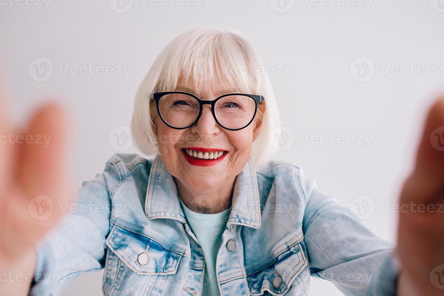 senior snygg kvinna med grått hår och i glasögon och jeansjacka kramar dig eller gör selfie. kärlek, kramar, anti age, positiva vibbar koncept foto