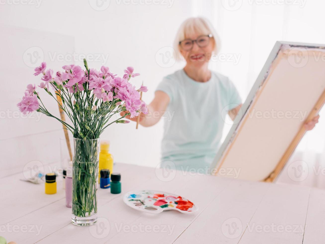 senior glad kvinna konstnär i glasögon med grått hår måla blommor i vas. kreativitet, konst, hobby, yrke koncept foto