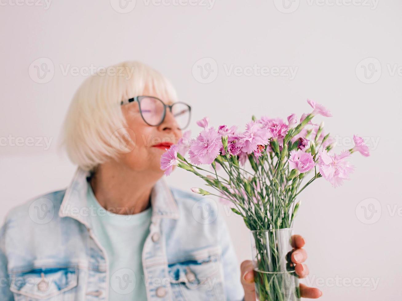 senior snygg kvinna i glasögon och jeansjacka luktar nejlikablommor i vas. blommor, pension, yrke, hobby koncept foto