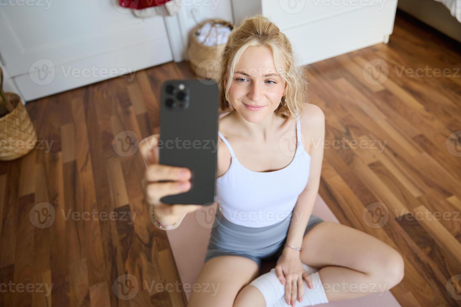 porträtt av skön ung kondition tränare, som visar Hem träna, tar selfie på mobil telefon medan Sammanträde på yoga sudd matta i sovrum, bär aktiva kläder foto