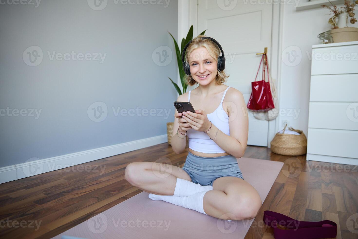 porträtt av ung sportig kvinna i trådlös hörlurar, Sammanträde med smartphone på sudd matta, använder sig av träna Träning app, kondition Ansökan på henne telefon, lyssnande i hörlurar foto