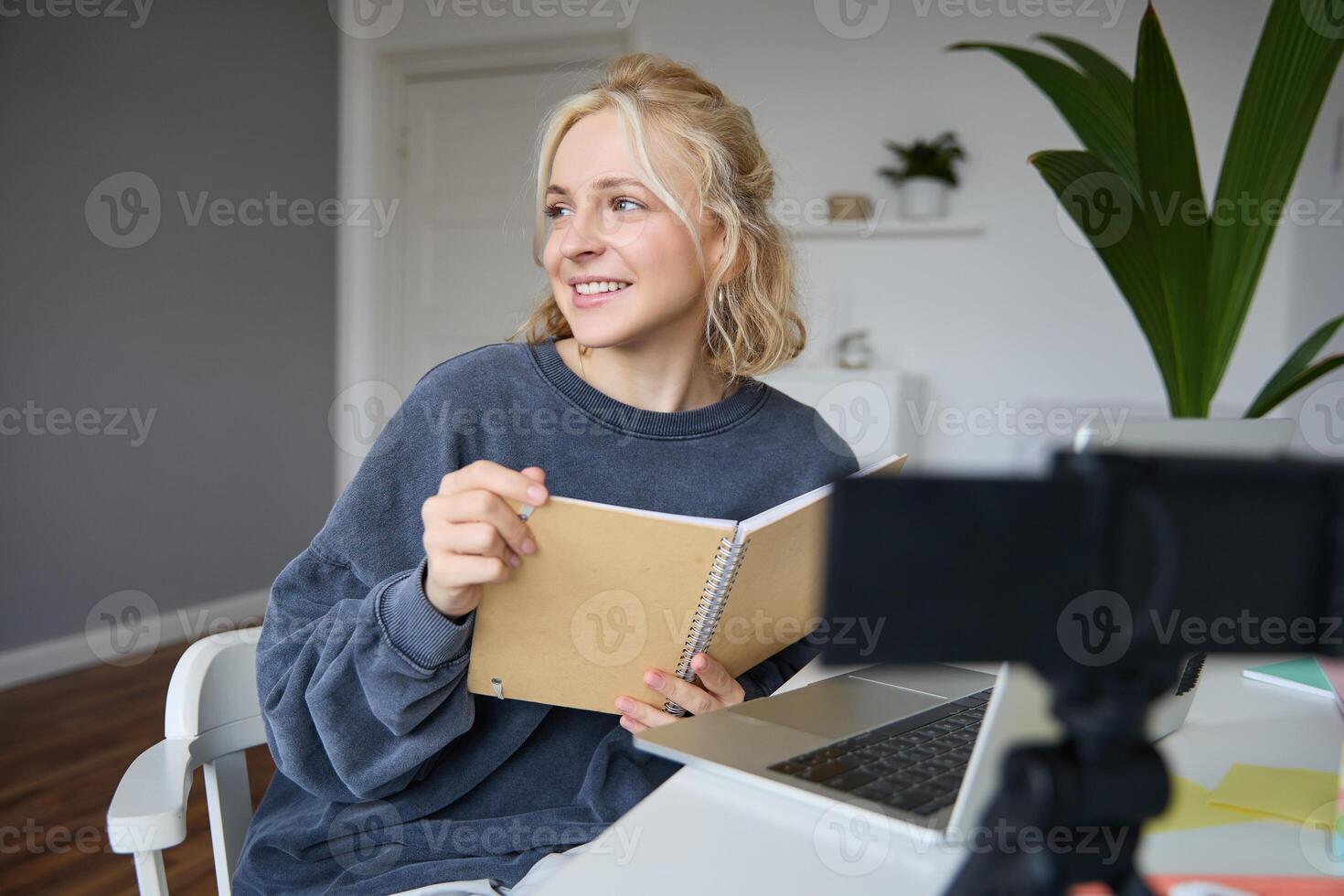 porträtt av leende blond kvinna, Sammanträde i sovrum, använder sig av bärbar dator och digital kamera, inspelning för livsstil blogg, läsning, använder sig av henne anteckningsbok foto
