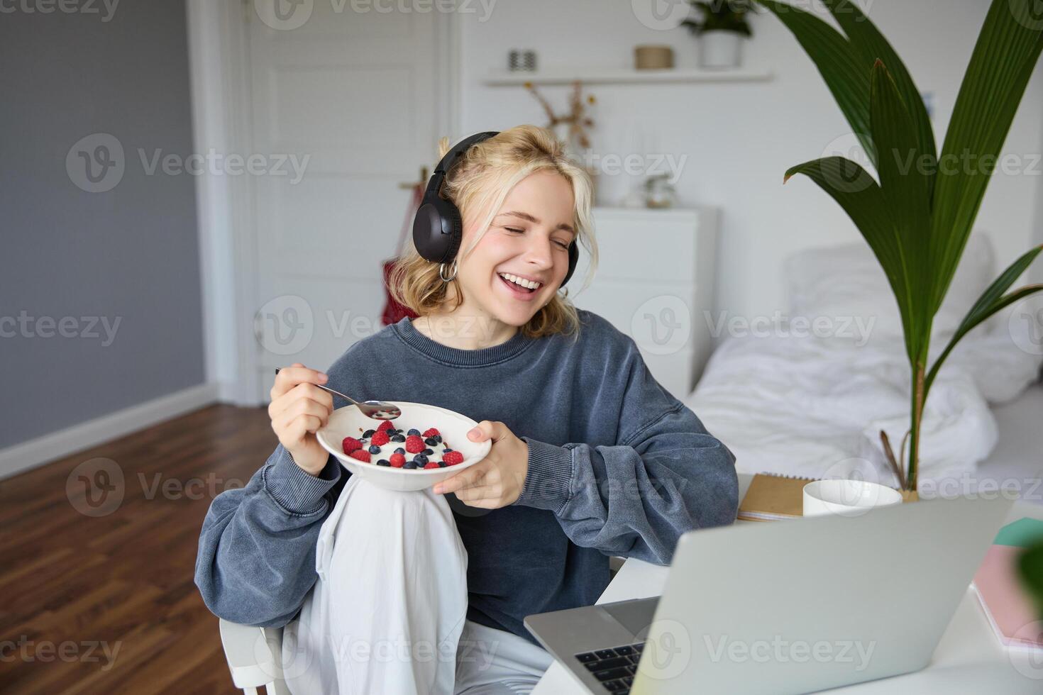 porträtt av leende ung kvinna, tittar på TV visa i hörlurar, äter frukost och ser på bärbar dator skärm foto