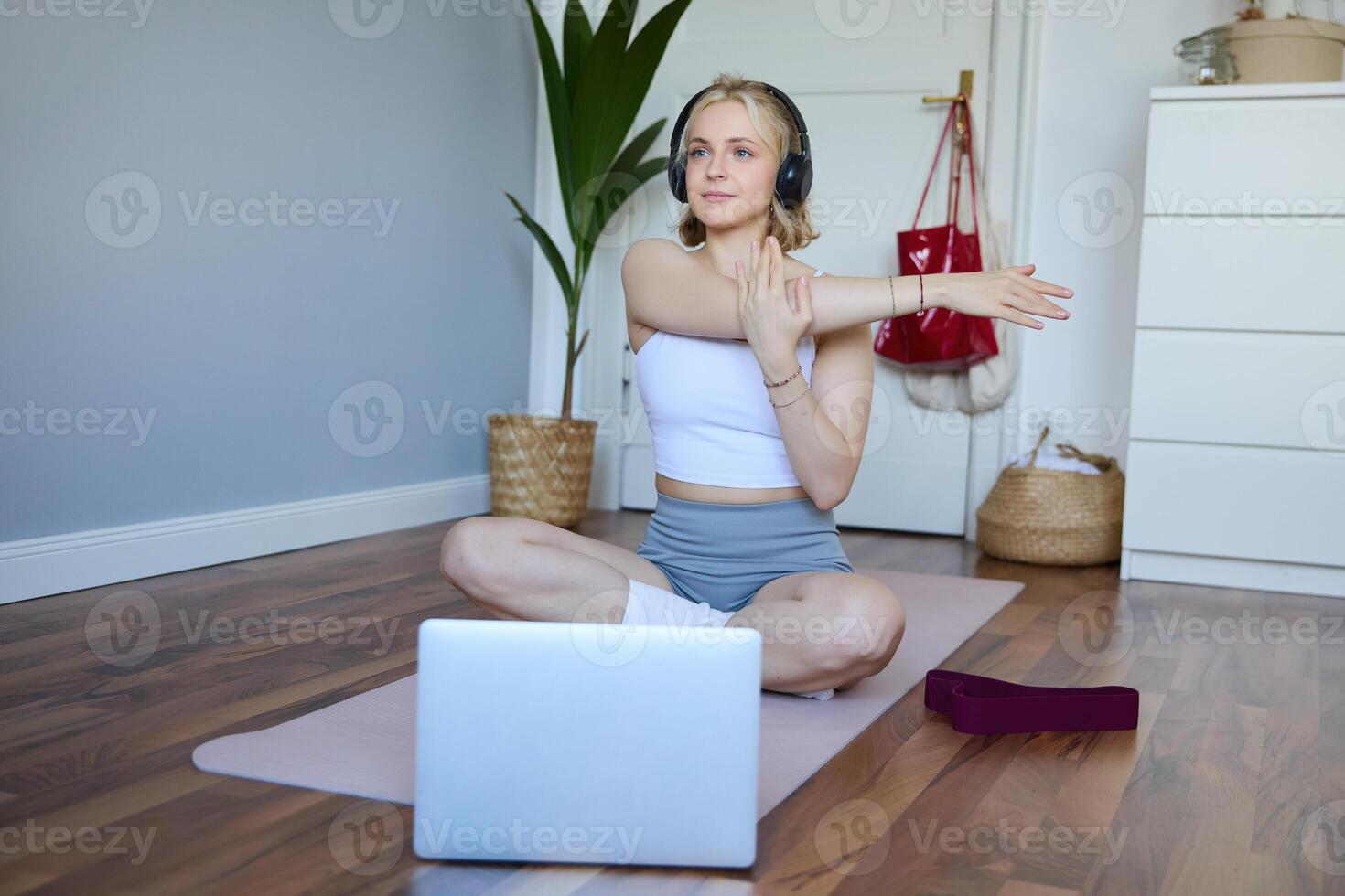 välbefinnande och kondition begrepp. kvinna i hörlurar, sitter på yoga matta, klockor träna på bärbar dator, sträckor vapen, gör övningar foto