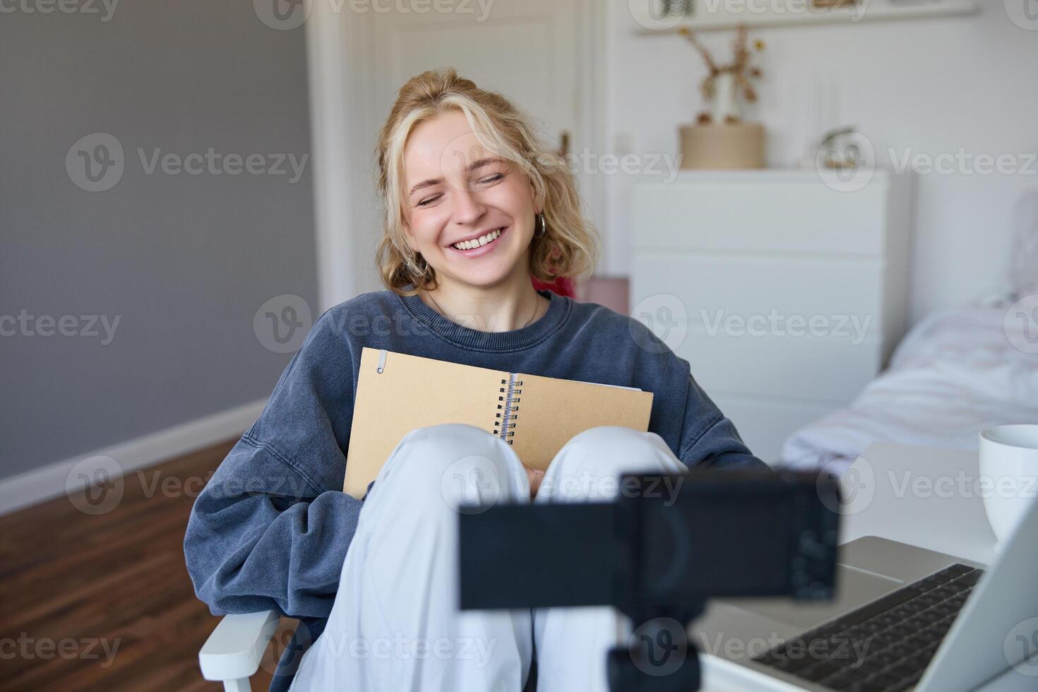 porträtt av blond leende kvinna, uppgifter på digital kamera på vilket sätt hon skrivningar i anteckningsbok, samtal till följare, håller på med livsstil blog innehåll i henne rum foto