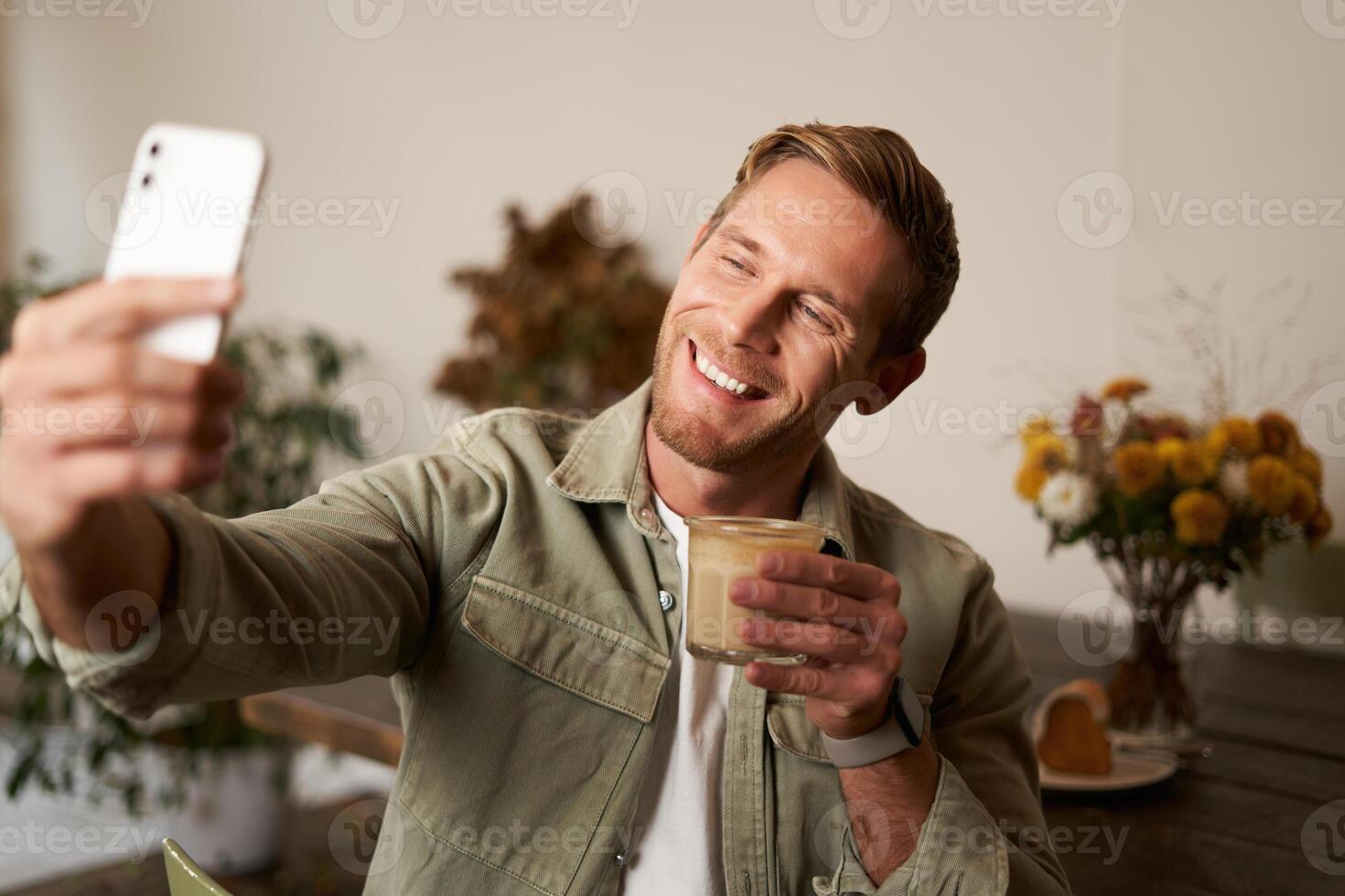 stänga upp porträtt av Lycklig, karismatisk ung blond man, tar selfie på smartphone, Framställ med kopp av kaffe, åtnjuter hans dryck i Kafé, rekommenderar en plats till hans följare uppkopplad foto
