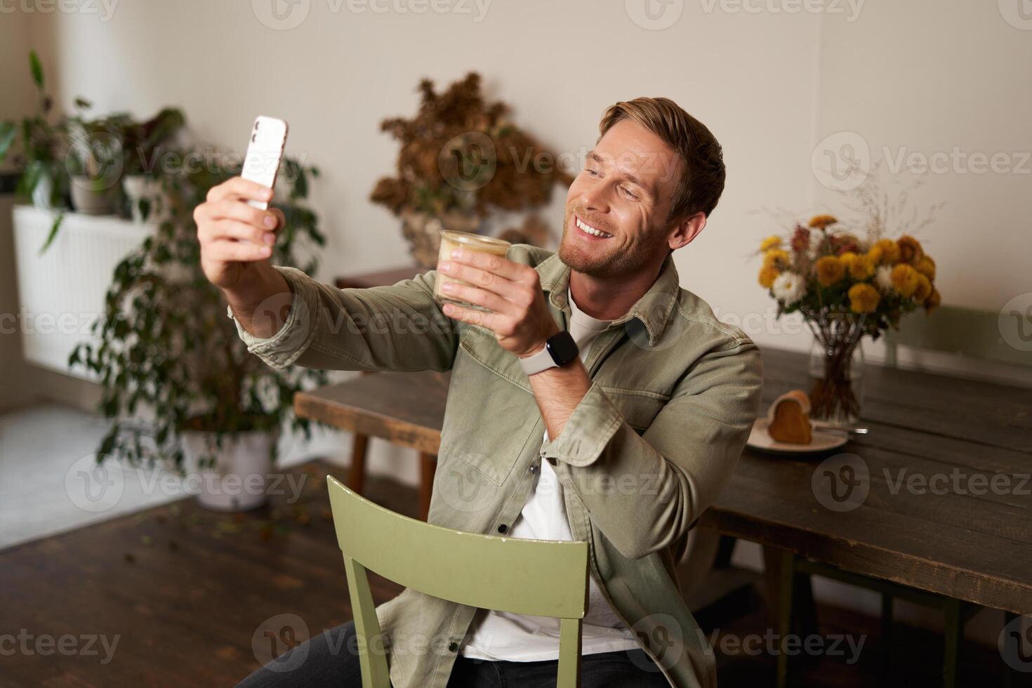 porträtt av ung stilig man tar slefie på hans mobil telefon app, sitter i Kafé med glas av kaffe, inlägg på social media Ansökan foto