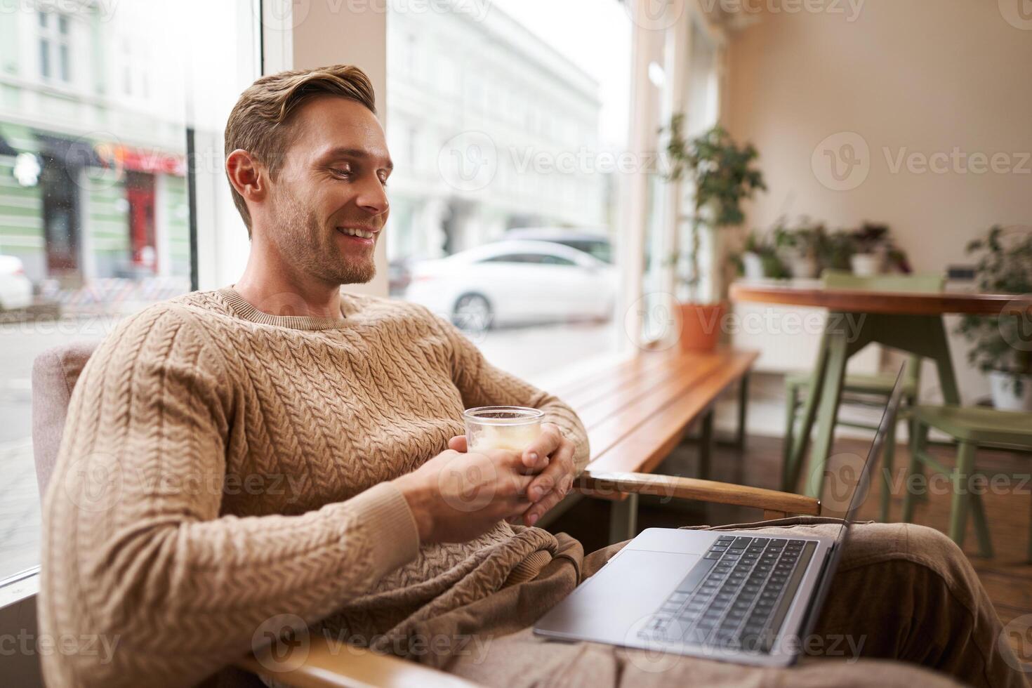 porträtt av stilig ung man sitter i Kafé, drycker kaffe och klockor på bärbar dator, ser på skärm med Lycklig leende, avkopplande i sam-arbetande Plats foto