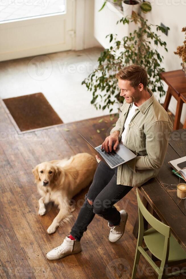 vertikal skott av stilig affärsman, ung man med bärbar dator lutar på tabell, Sammanträde på sam-arbetande Plats med hans hund. ung försäljare checkar hans meddelanden, leende, arbetssätt i ett kontor med en sällskapsdjur foto