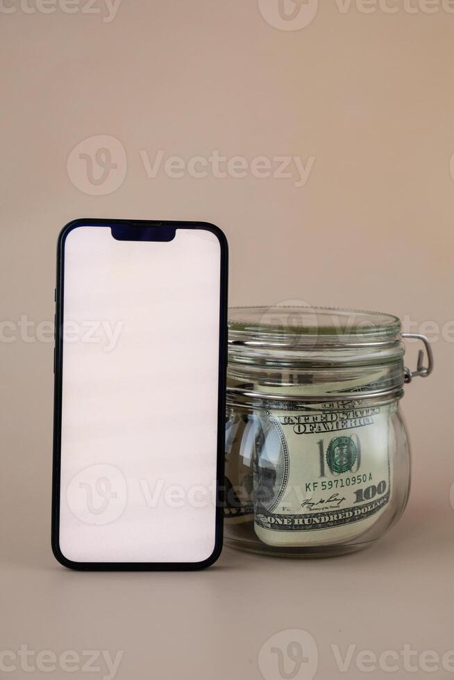 vit smartphone skärm falsk upp mall i vertikal placera på beige bakgrund. kopia Plats app hemsida reklam. burk fylld med dollar kontanter. begrepp av mobil Ansökan och teknologi företag besparingar. foto