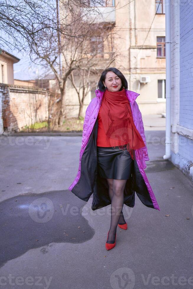 en eleganta kvinna i en lång ljus rosa, magenta täcka och röd skor promenader de gator av de stad foto
