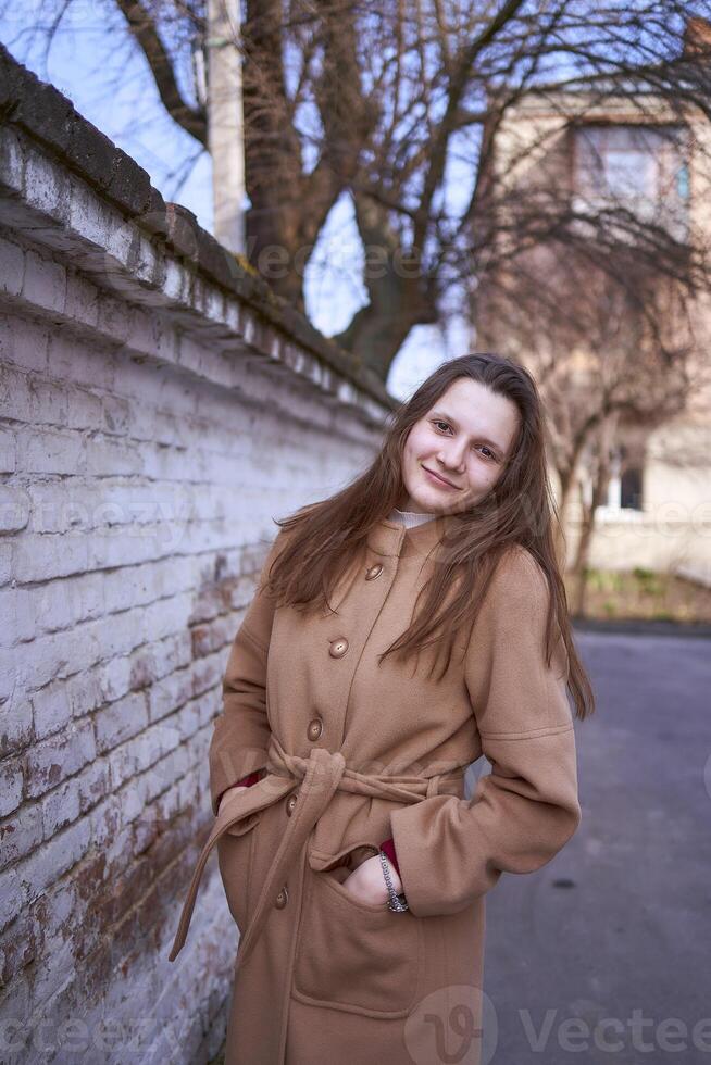 en flicka i en brun täcka är Lycklig nära en vit vägg på en vår dag foto