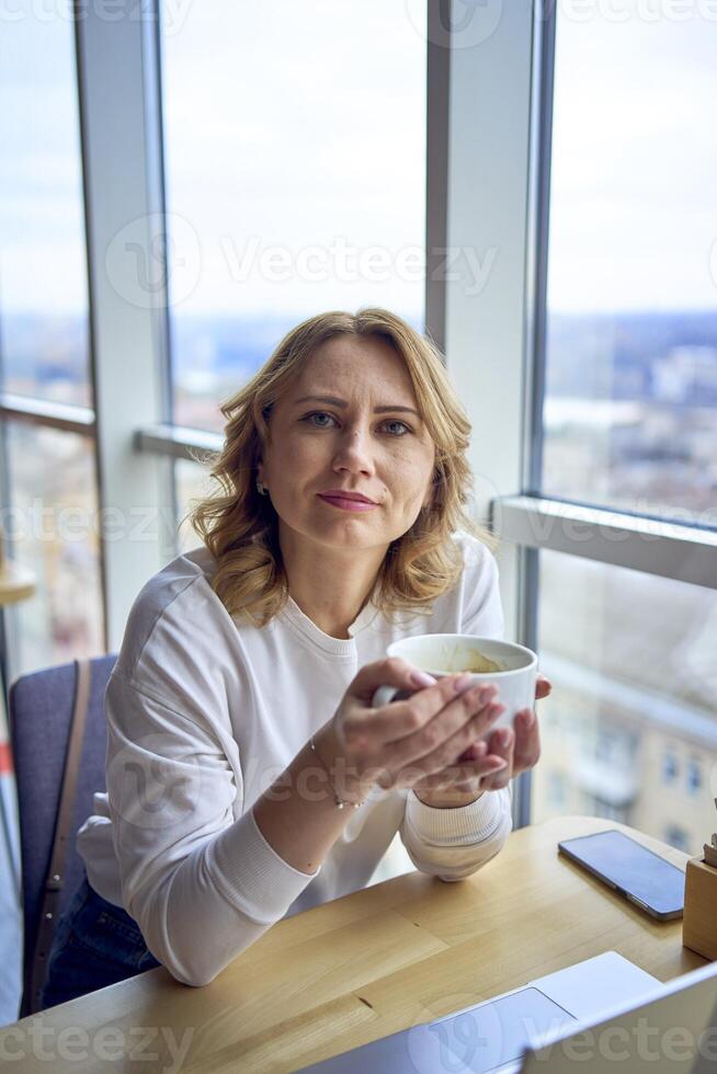 mitten ålder kvinna med blond hår dricka kaffe och arbetssätt på en bärbar dator i en coworking Plats med panorama- fönster och en se av de stad från ovan foto