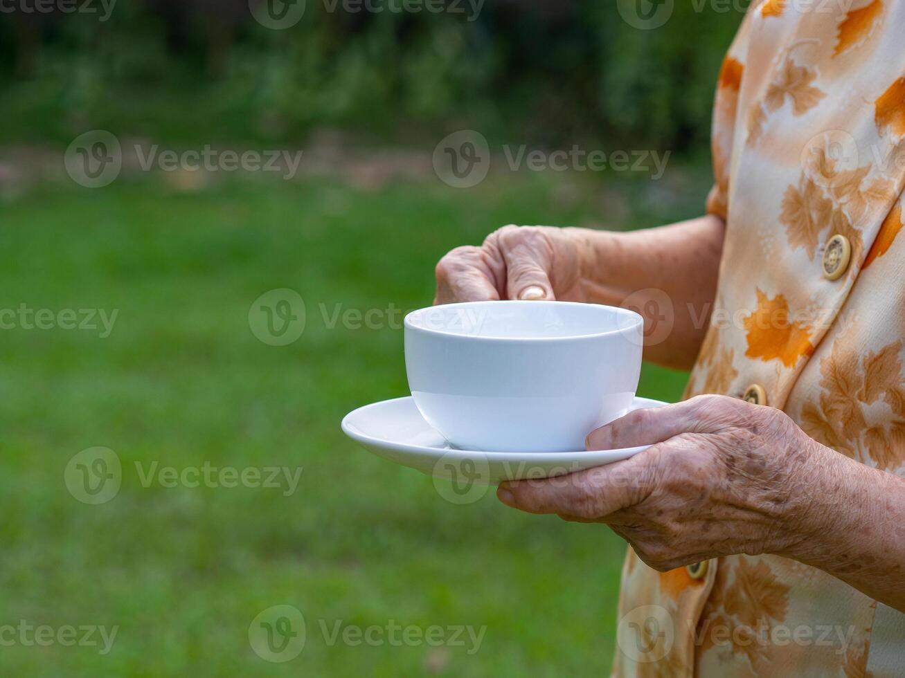 närbild av senior kvinna händer innehav en vit kopp medan stående i en trädgård med kopia Plats för text. begrepp av åldrig människor och avslappning foto