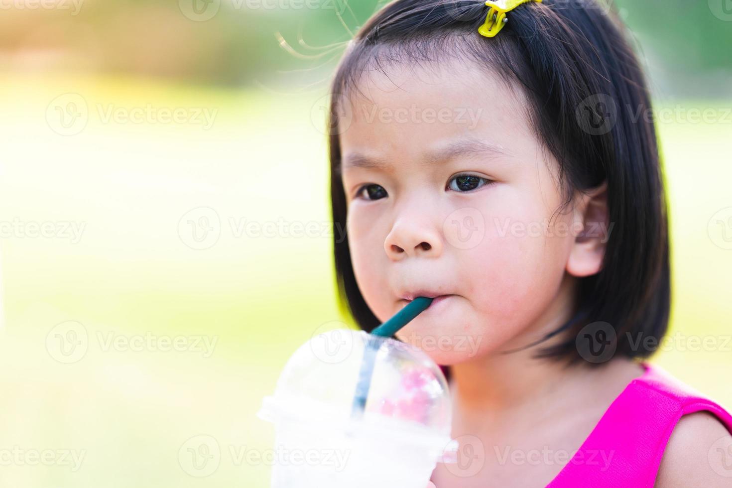 söt asiatisk tjej suger vatten från plastglas med grönt halm. en varm dag ute. huvudskott. foto