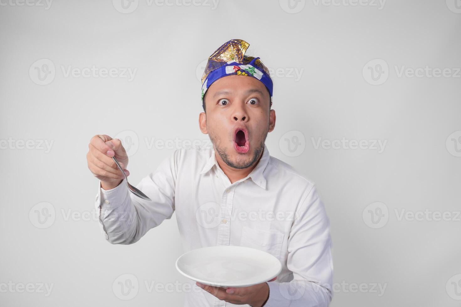 chockade ung balinesisk man i vit skjorta och traditionell huvudbonad innehav ett tömma tallrik med kopia Plats och sked, känsla överraskad förbi de mat meny foto