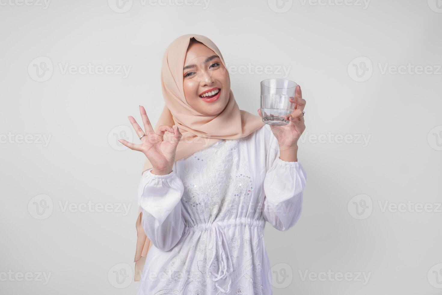 ung asiatisk muslim kvinna i vit klänning och hijab innehav och dricka kopp av färsk vatten medan framställning ett Okej, Bra, trevlig tecken gest använder sig av fingrar foto