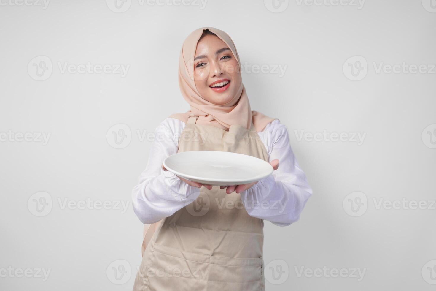 skön ung asiatisk muslim kvinna i hijab och grädde förkläde presenter ett tömma tallrik med kopia Plats över Det, som visar de mat meny för iftar foto