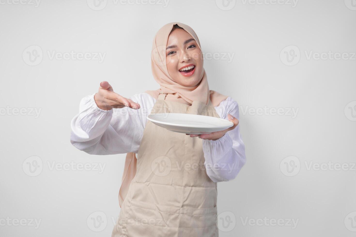 porträtt av en glad ung asiatisk muslim kvinna i en slöja hijab och grädde förkläde pekande till ett tömma tallrik med kopia Plats över Det, presenter mat meny foto