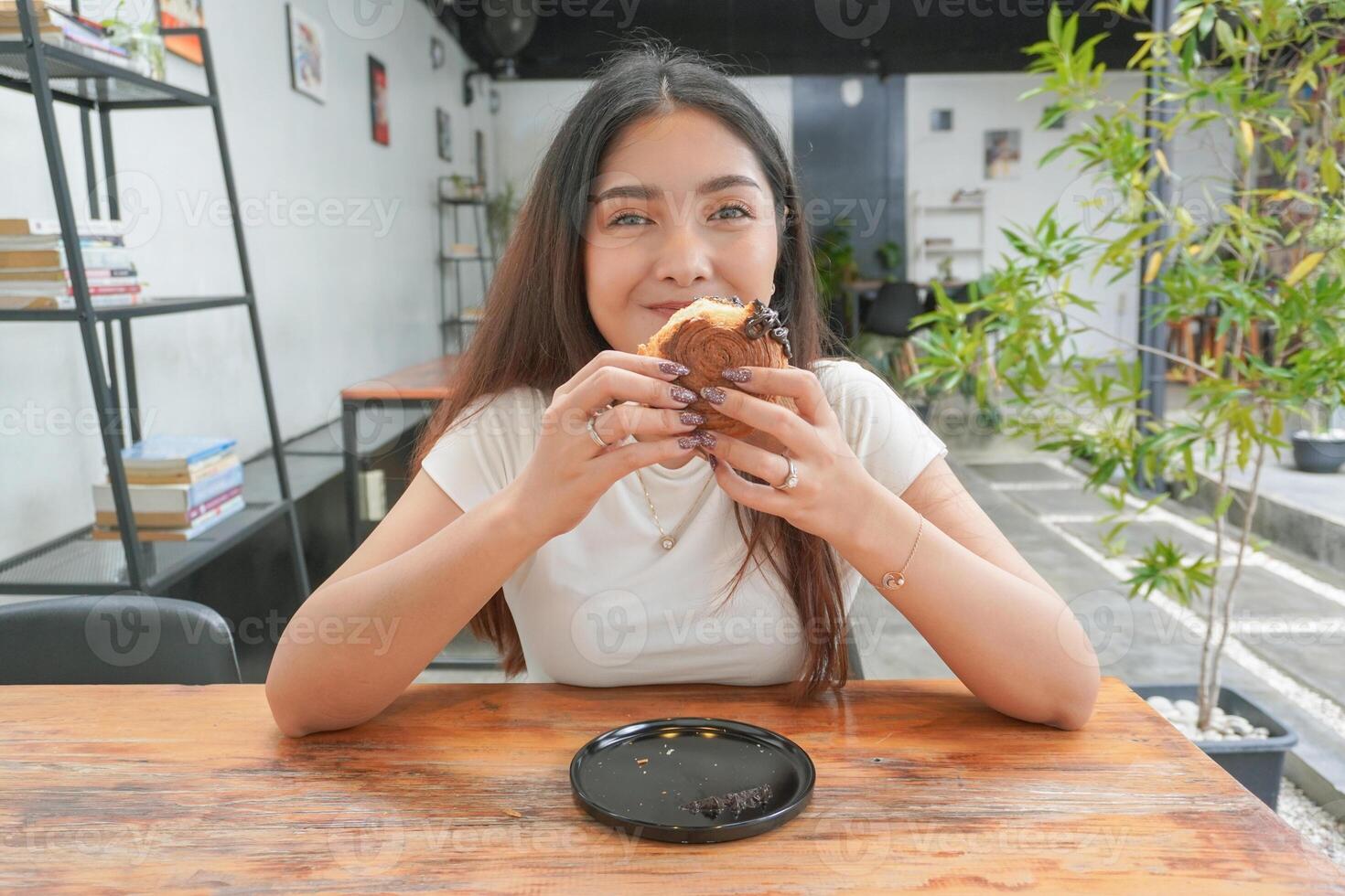 skön asiatisk ung kvinna äter utsökt bakverk som heter cromboloni på en kafé. de lady biter bit av choklad fyllning cromboloni, ser Lycklig på de Kafé foto