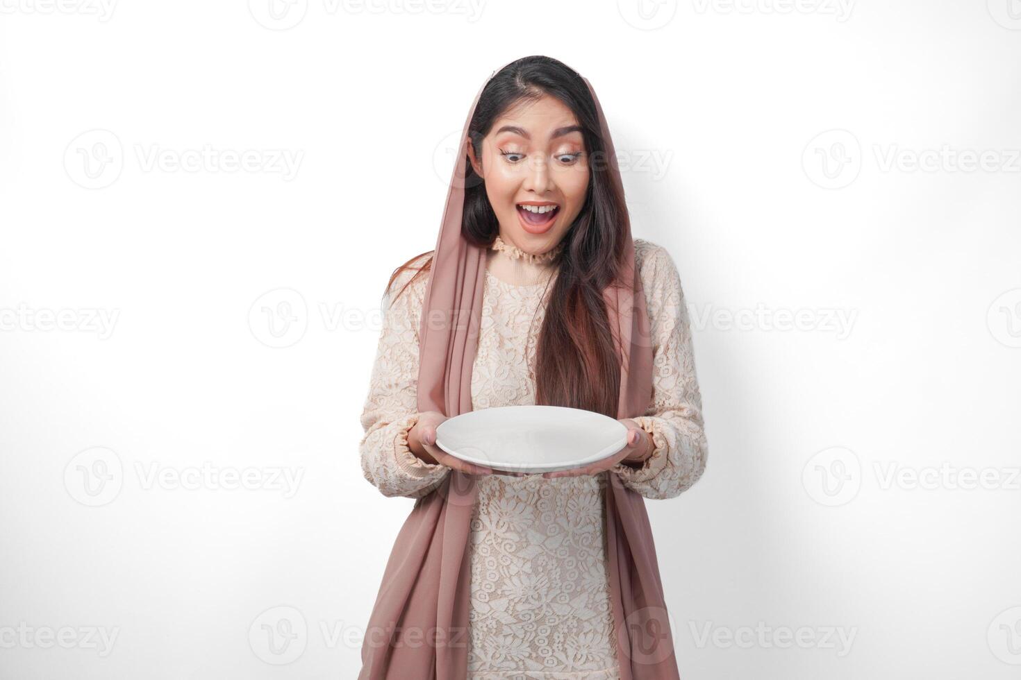överraskad asiatisk muslim kvinna i slöja slöja hijab innehav tallrik med kopia Plats inuti, isolerat vit bakgrund foto
