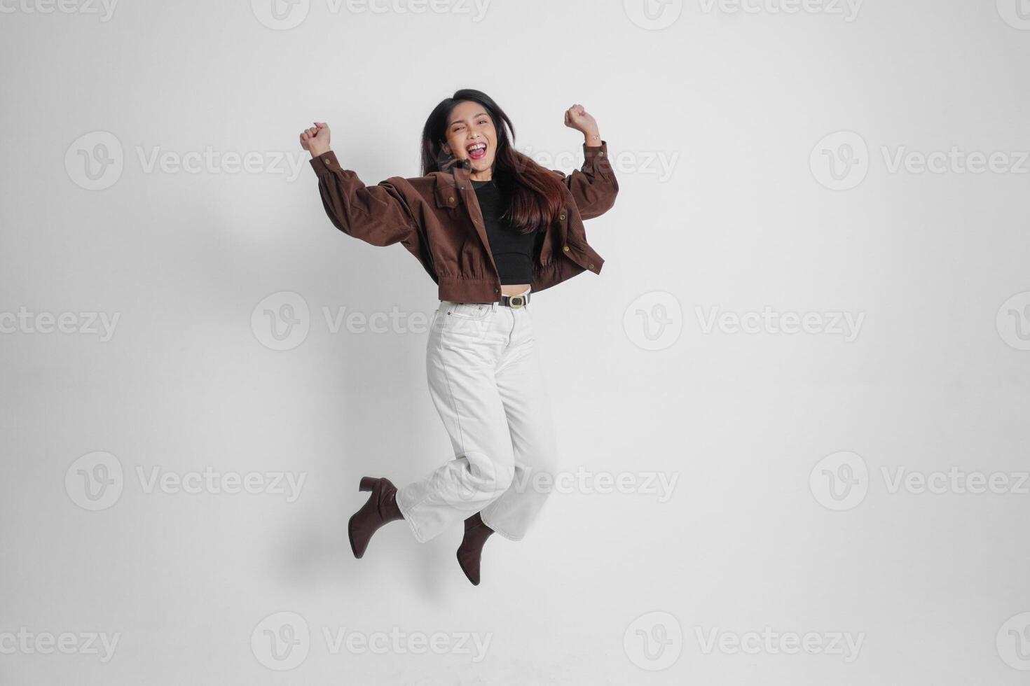 aktiva asiatisk flicka är Hoppar och höjning händer upp ha på sig brun jacka, isolerat vit Färg bakgrund foto