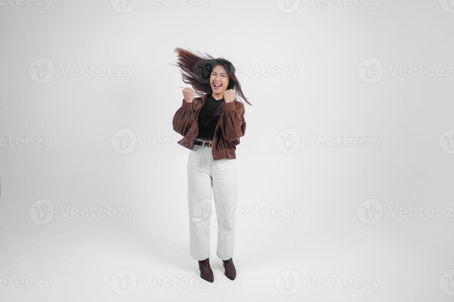 aktiva asiatisk flicka är Hoppar och höjning händer upp ha på sig brun jacka, isolerat vit Färg bakgrund foto