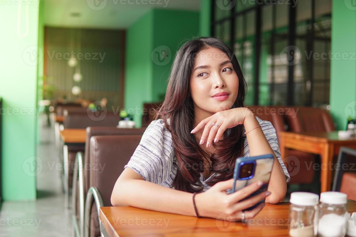 dagdrömma ung asiatisk kvinna bär klänning Sammanträde på en restaurang för en frukost, innehav en smartphone medan stöttning henne haka förlorat i trodde foto