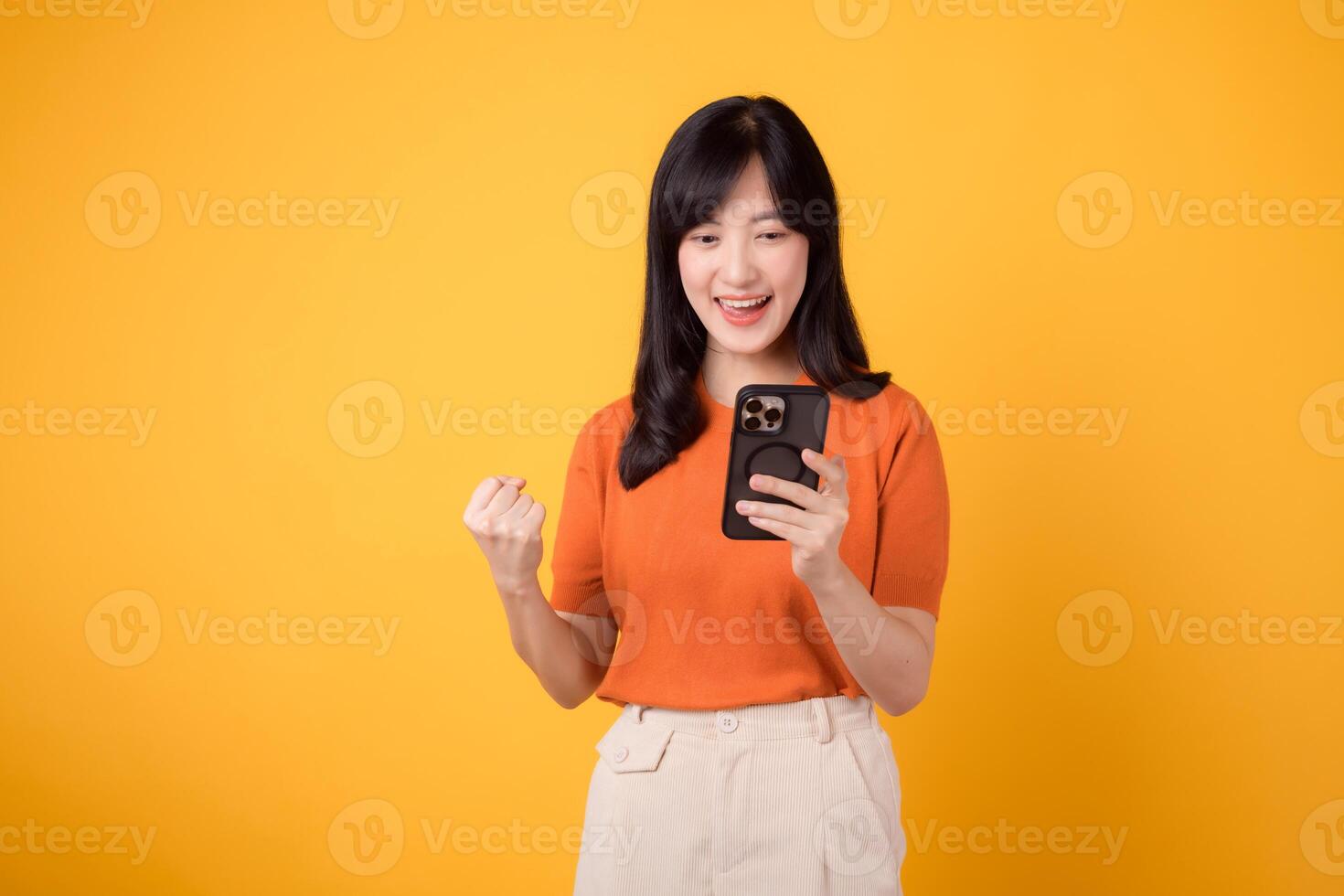 vibrerande asiatisk kvinna i 30-talet, bär orange skjorta, använder sig av smartphone med näve upp hand tecken på vibrerande gul bakgrund. fira ny app begrepp. foto
