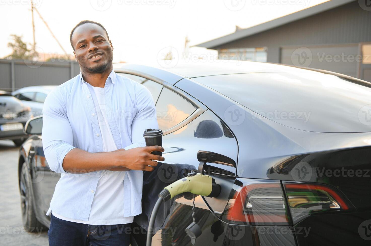 eleganta afrikansk man med coffe kopp i hand skär plugg in i de elektrisk bil laddning uttag foto