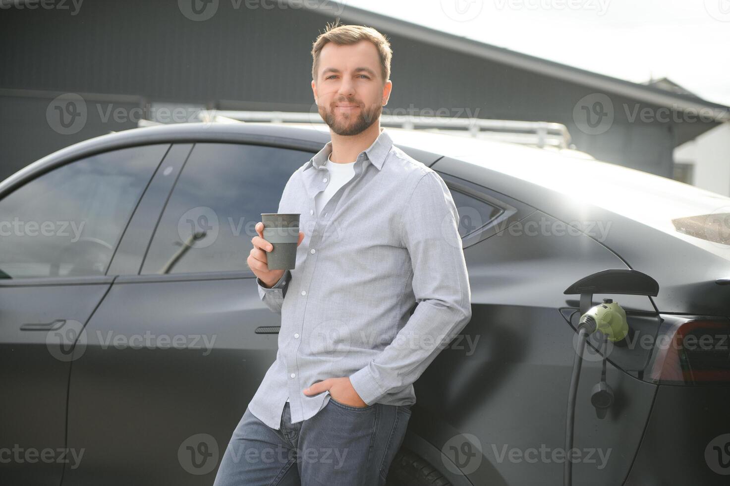 stilig skäggig man i tillfällig ha på sig, stående på de laddning station och laddare för ett elektrisk bil. eco elektrisk bil begrepp foto