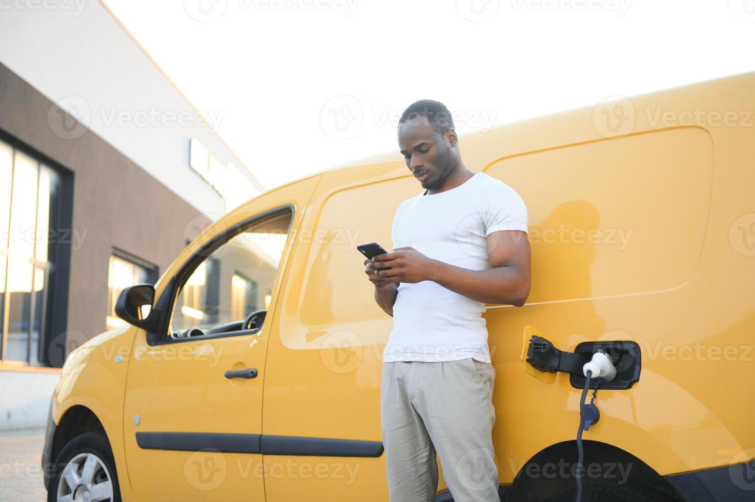 en afrikansk amerikan man står Nästa till gul elektrisk leverans skåpbil på elektrisk fordon laddning stationer foto