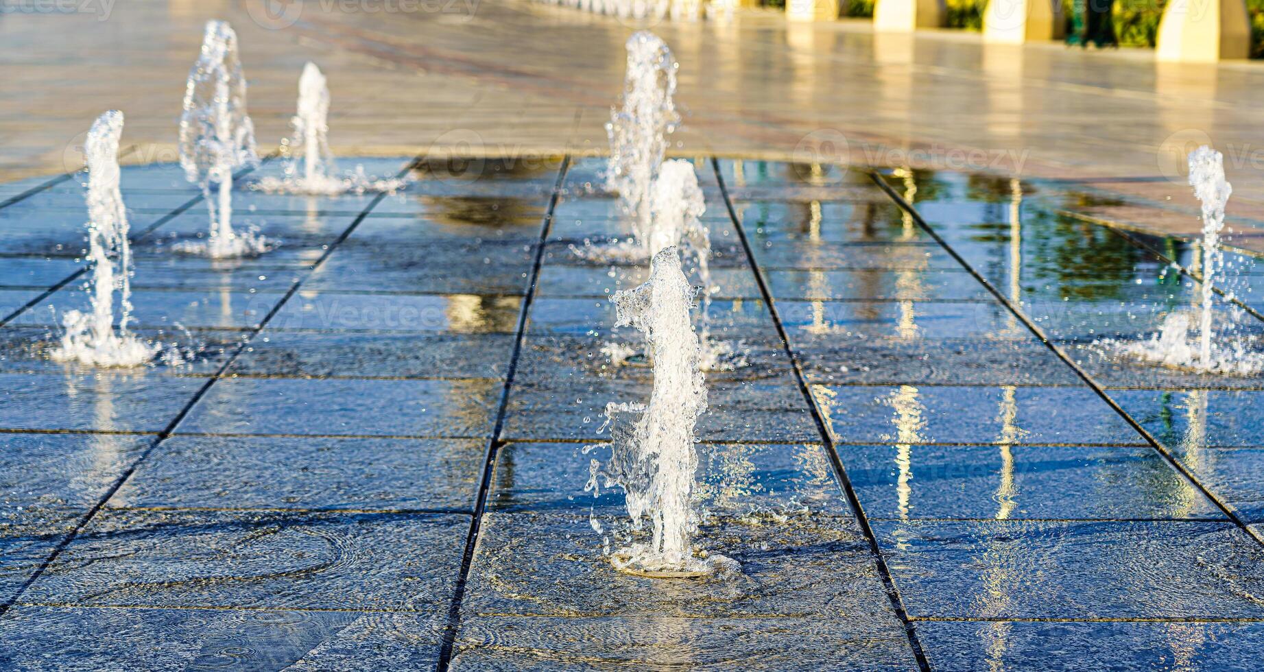strömmar av stänk små fontäner på våt stenläggning plattor, upplyst förbi de Sol i en sommar. foto