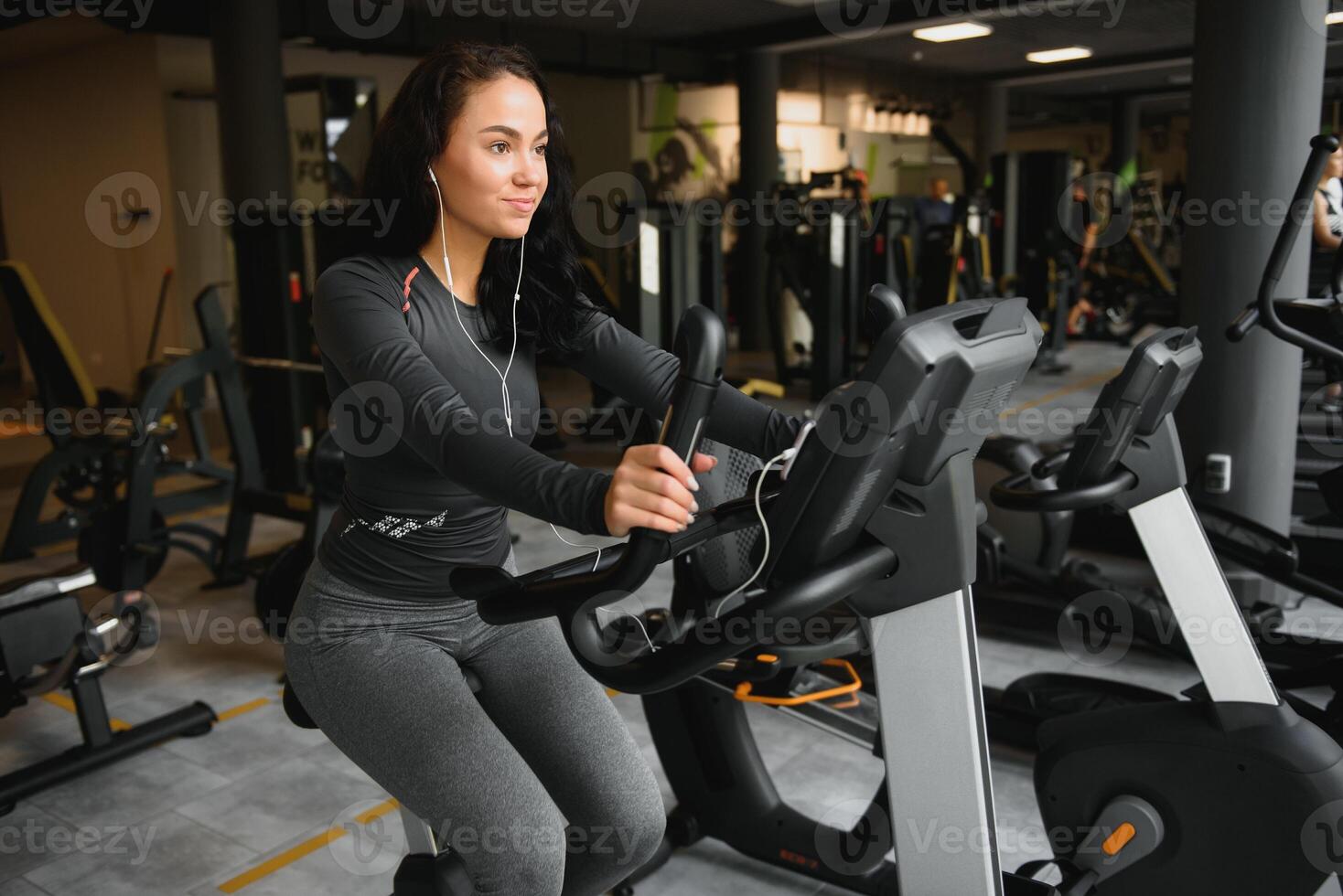 ung kvinna med hörlurar håller på med övningar på brevpapper cykel i en Gym eller kondition Centrum. ung sportig kvinna i Gym lyssna musik från smartphone. kvinnor håller på med konditionsträning övningar foto