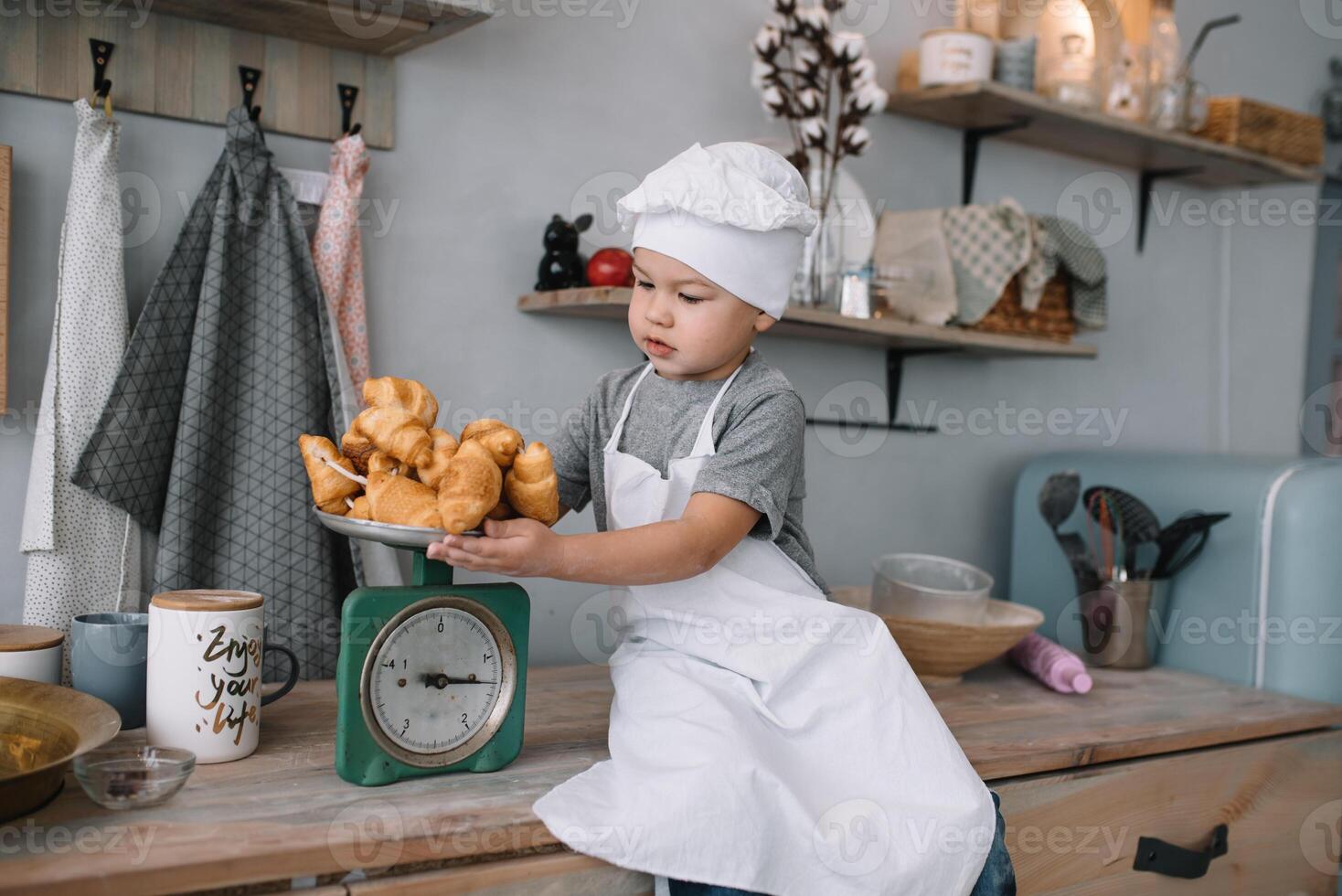 ung pojke söt på de kök laga mat kock i vit enhetlig och hatt nära tabell. hemlagad pepparkaka. de pojke kokta de choklad småkakor. foto