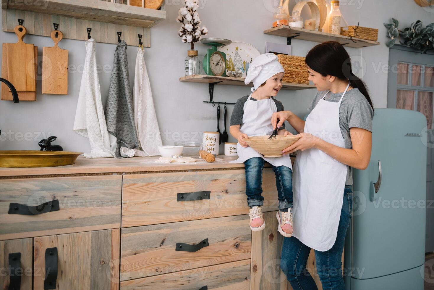 ung Lycklig mamma och henne bebis laga mat småkakor på Hem i de kök. jul hemlagad pepparkaka. söt pojke med mor i vit enhetlig och hatt kokta choklad småkakor foto