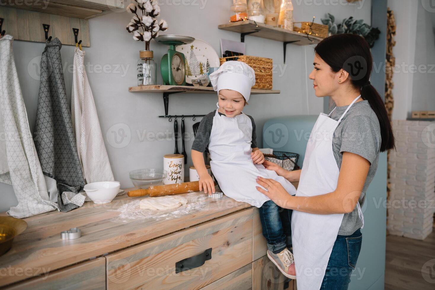 ung Lycklig mamma och henne bebis laga mat småkakor på Hem i de kök. jul hemlagad pepparkaka. söt pojke med mor i vit enhetlig och hatt kokta choklad småkakor. foto