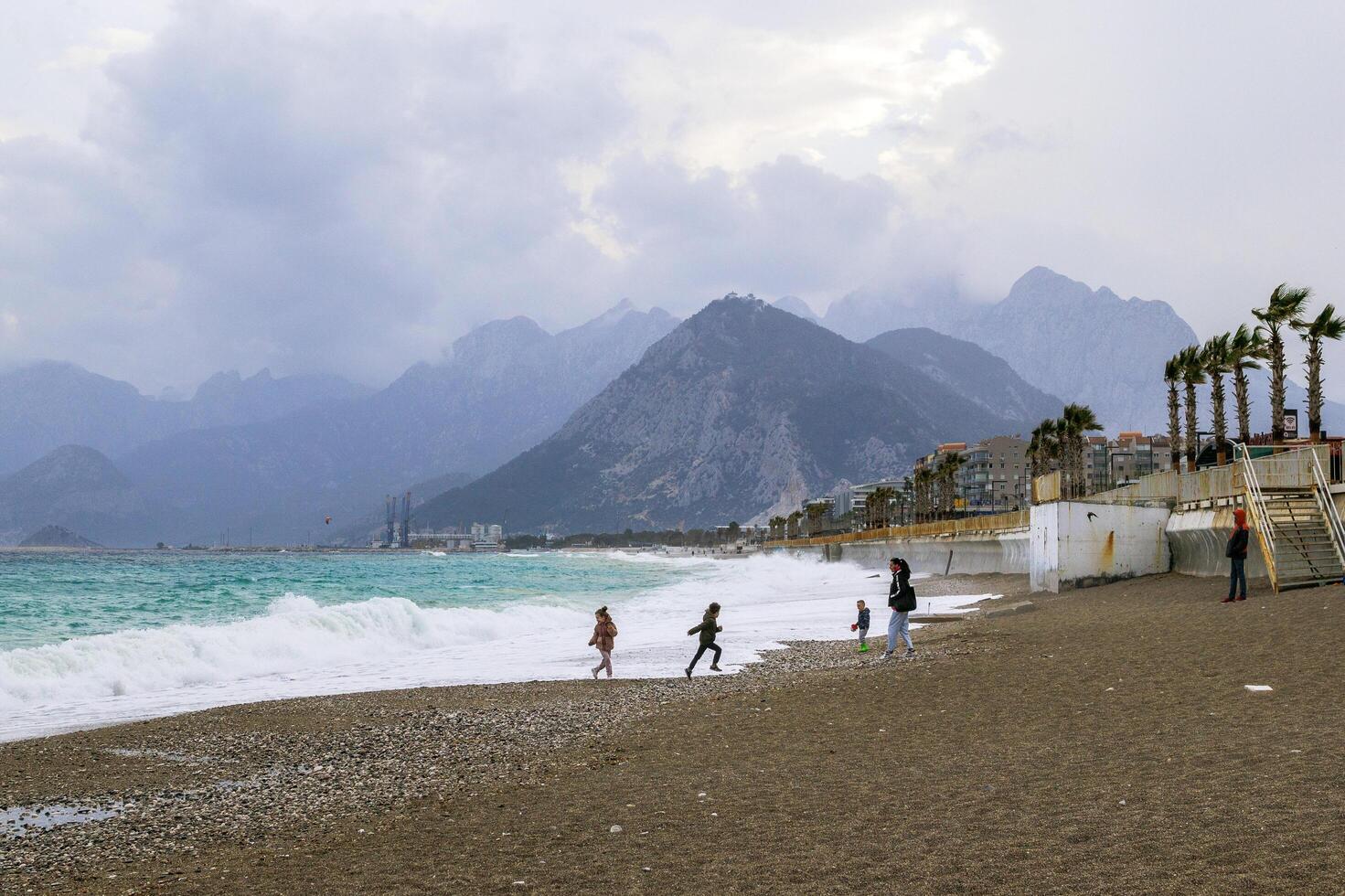 antalya, Kalkon - Mars 05, 2023. oigenkännlig människor är gående och spelar på vägbank nära till stormig hav med stor vågor och bergen på bakgrund. foto