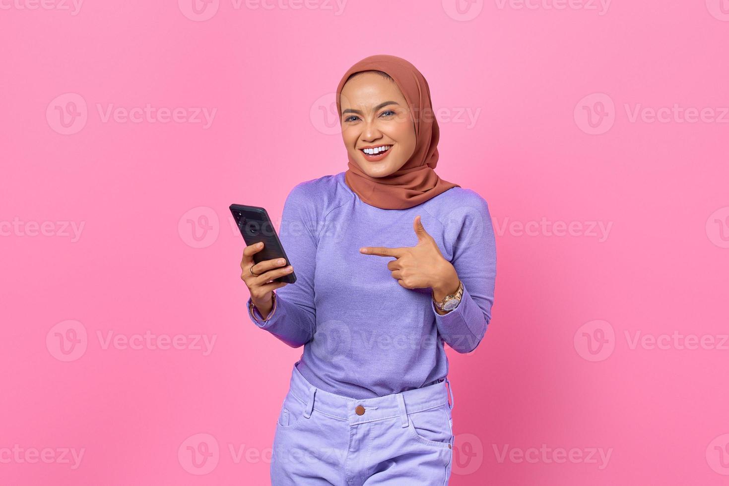 porträtt av leende ung asiatisk kvinna pekar finger på en mobiltelefon på rosa bakgrund foto