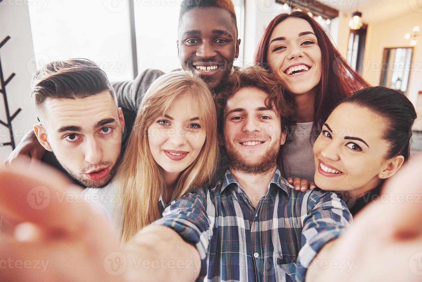 vänner har kul på restaurang. tre pojkar och tre tjejer gör selfie och skrattar. på förgrunden pojke håller smart telefon. alla bär vardagskläder foto