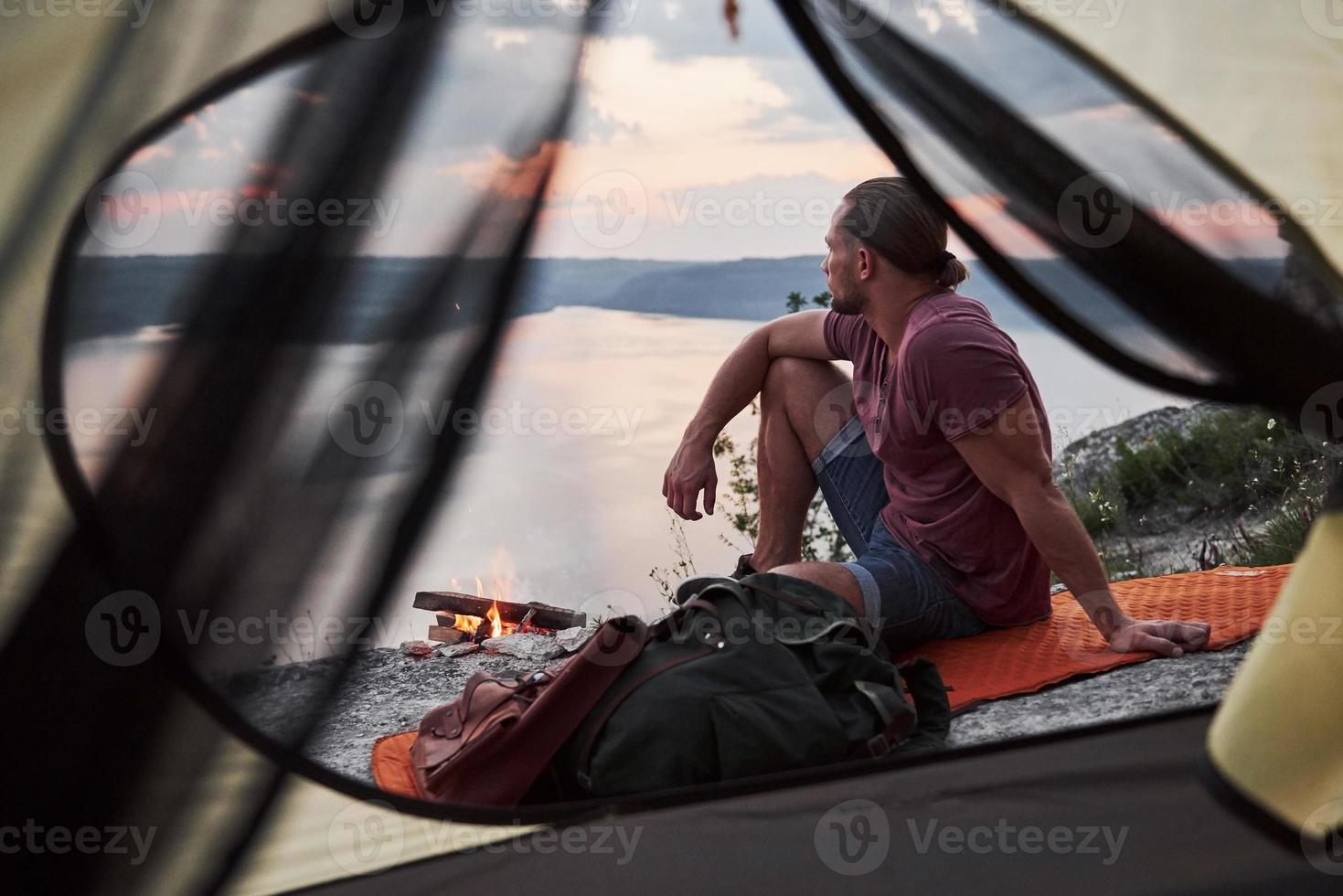 utsikt från resenärens tält med ryggsäck som sitter på toppen av berget och njuter av utsikten över en flod eller sjö. resa längs berg och kust, frihet och aktivt livsstilskoncept foto