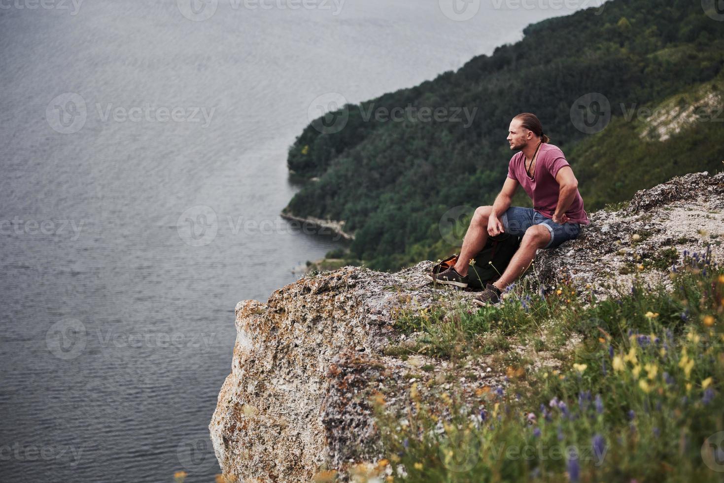 resenär med ryggsäck som sitter på toppen av berget och njuter av utsikten över vattenytan. resa längs berg och kust, frihet och aktivt livsstilskoncept foto