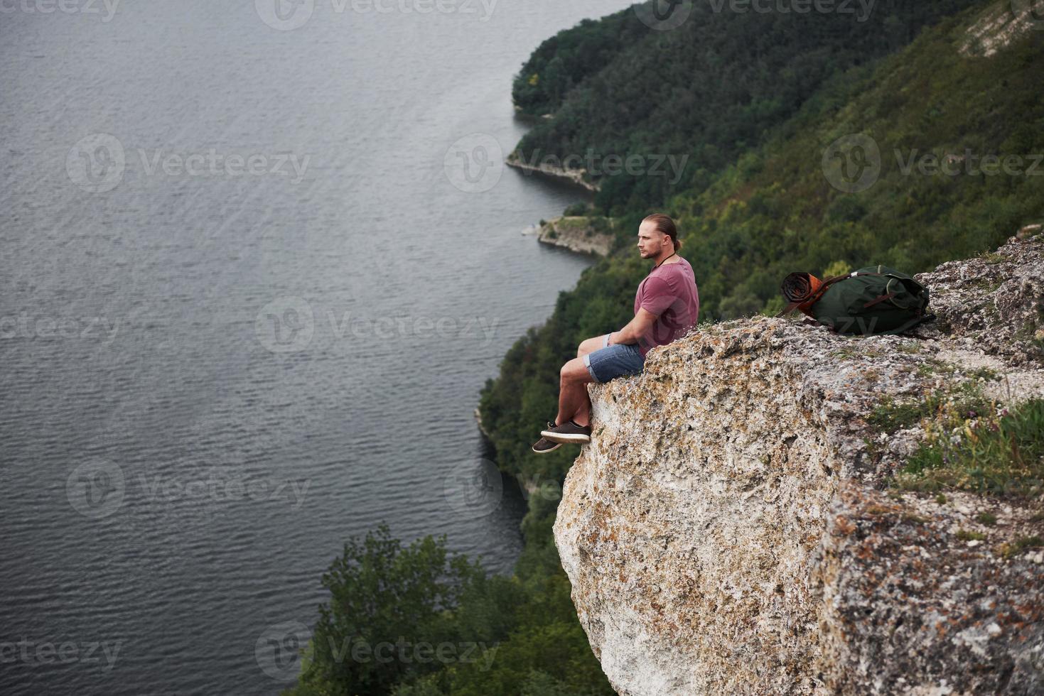 resenär med ryggsäck som sitter på toppen av berget och njuter av utsikten över vattenytan. resa längs berg och kust, frihet och aktivt livsstilskoncept foto