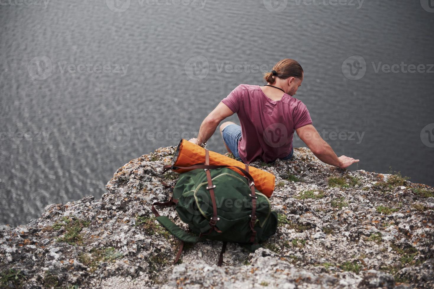 resenär med ryggsäck som sitter på toppen av berget och njuter av utsikten över vattenytan. resa längs berg och kust, frihet och aktivt livsstilskoncept foto