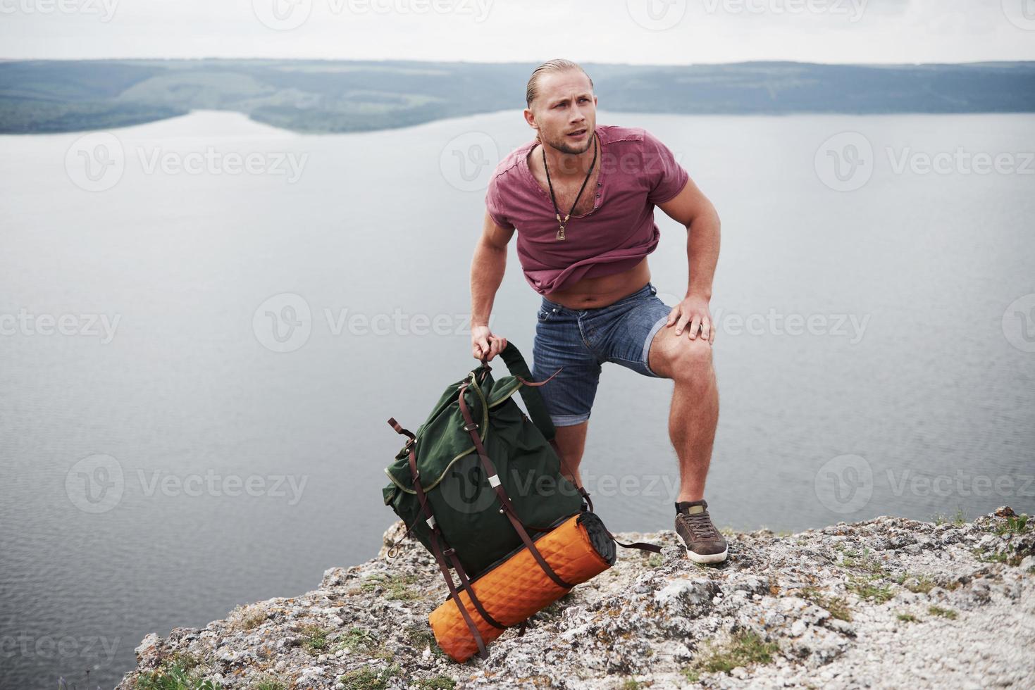 resenär med ryggsäck som sitter på toppen av berget och njuter av utsikten över vattenytan. resa längs berg och kust, frihet och aktivt livsstilskoncept foto